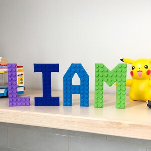 May include: The name "LIAM" spelled out in colorful plastic letters. Each letter is made of LEGO bricks. The letters are arranged on a light wood shelf.