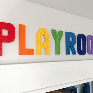 May include: Colorful "PLAYROOM" letters in red, orange, yellow, green, blue, purple, and pink are mounted on a white wall. The letters are designed to resemble toy building blocks, adding a playful touch to the room's decor.