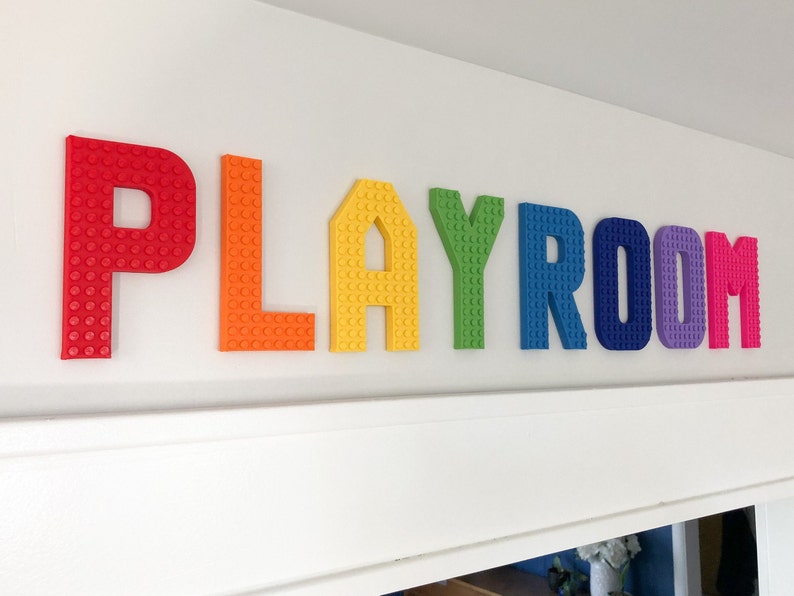 May include: Colorful "PLAYROOM" letters in red, orange, yellow, green, blue, purple, and pink are mounted on a white wall. The letters are designed to resemble toy building blocks, adding a playful touch to the room's decor.
