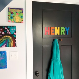 May include: A black door with the name "HENRY" spelled out in colorful block letters. A teal hooded jacket hangs from a hook below the name. Artwork hangs on the white wall to the left.