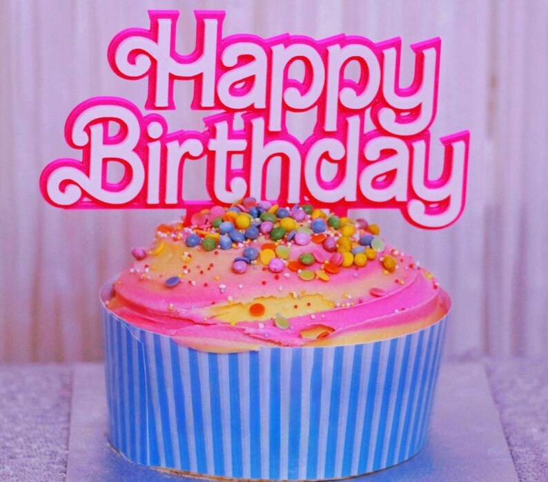 May include: A frosted cupcake with pink and yellow icing, topped with colorful sprinkles and a pink and white "Happy Birthday" cake topper. The cupcake is in a blue and white striped liner.