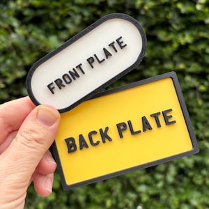 Puede incluir: Dos placas rectangulares con el texto "FRONT PLATE" y "BACK PLATE". La placa frontal es blanca con borde negro, y la placa trasera es amarilla con borde negro. El texto es negro.