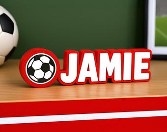 May include: A red nameplate with the name "JAMIE" in white, with a soccer ball graphic. A soccer ball is visible in the background. The nameplate is on a wooden surface. The nameplate is a decorative item.