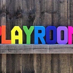 May include: Colorful, rainbow-colored letters spelling "PLAYROOM" are arranged on a wooden shelf against a weathered wood background. The letters have a textured, brick-like appearance, adding a playful touch to the room's decor.