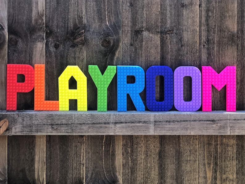 May include: Colorful, rainbow-colored letters spelling "PLAYROOM" are arranged on a wooden shelf against a weathered wood background. The letters have a textured, brick-like appearance, adding a playful touch to the room's decor.