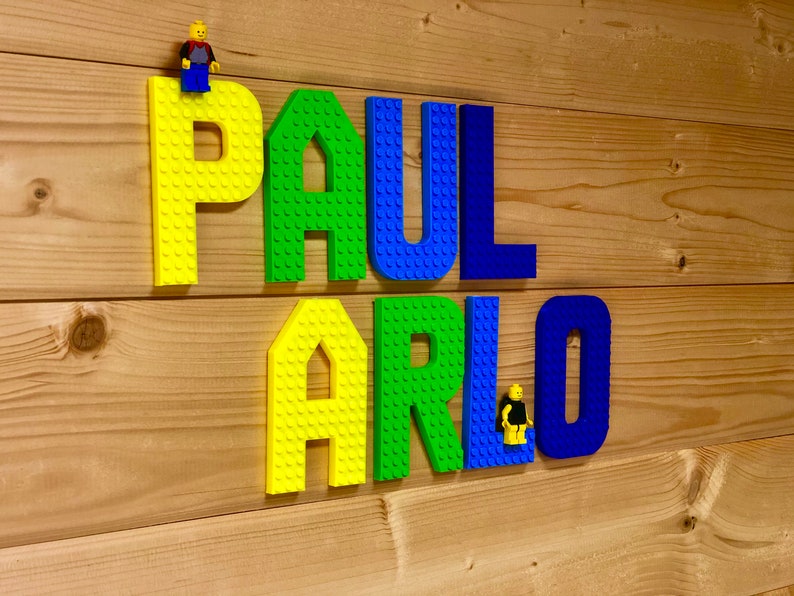 May include: Colorful LEGO-style letters spelling "PAUL ARLO" are mounted on a wooden plank background. The letters are yellow, green, and blue. Small LEGO figures are placed on top of the letters.