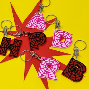 May include: Set of six keychains featuring different letters of the alphabet. Each letter is made of black and white plastic with a spiderweb design. The keychains are attached to silver metal rings.