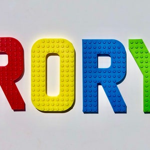 May include: Colorful letters spelling "RORY" in red, yellow, blue, and green. The letters are designed to resemble toy building blocks, with a textured surface of small circular studs. The letters are arranged on a white background.