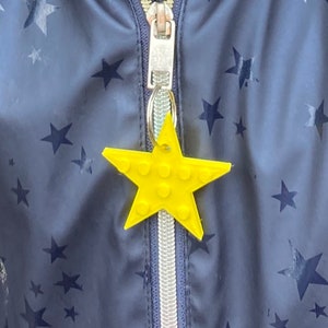 Könnte beinhalten: Eine blaue Jacke mit einem Reißverschluss und einem gelben sternförmigen Schlüsselanhänger. Die Jacke hat einen weißen Reißverschluss und ein weißes Futter. Die Jacke ist mit kleinen schwarzen Sternen bedeckt.