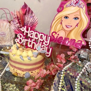 May include: A birthday cake with a mermaid tail and seashell decorations. The cake is topped with a pink "Happy Birthday" sign and a Barbie-themed cutout. The cake is surrounded by pink flowers and decorative elements.