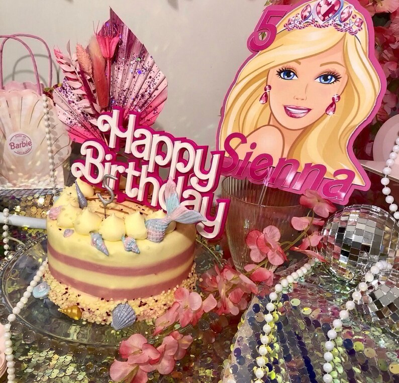 May include: A birthday cake with a mermaid tail and seashell decorations. The cake is topped with a pink "Happy Birthday" sign and a Barbie-themed cutout. The cake is surrounded by pink flowers and decorative elements.