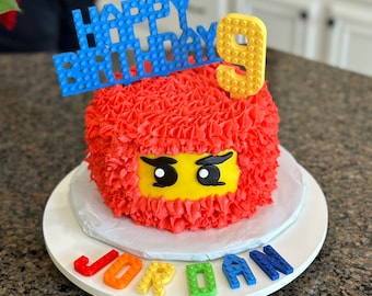 Op de afbeelding: Een rode en gele Lego-thema verjaardagstaart met een blauwe Lego 'Happy Birthday' topper en een gele Lego '9' erop. De taart is versierd met rode frosting en een Lego gezicht. De taart staat op een witte taartstandaard met de naam 'Jordan' gespeld in Lego-stenen.