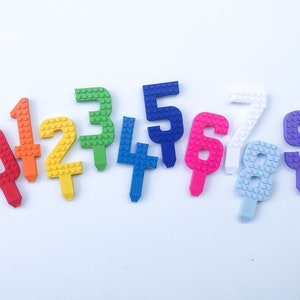 May include: A set of ten colorful plastic number candles, each shaped like a Lego brick. The numbers are 0 through 9, and the colors are red, orange, yellow, green, blue, purple, pink, white, and light blue.