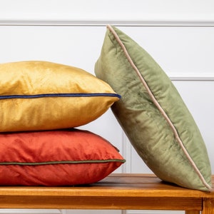 May include: Three decorative throw pillows stacked on a wooden bench. The top pillow is a muted green with a light pink trim. The middle pillow is a burnt orange with a dark green trim. The bottom pillow is a mustard yellow with a dark blue trim.