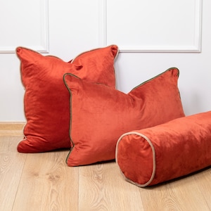 Pu&ograve; includere: Tre cuscini da lancio in velluto color ruggine con bordino crema e verde. I cuscini sono disposti su un pavimento in legno chiaro. Un cuscino &egrave; un cuscino a rullo.