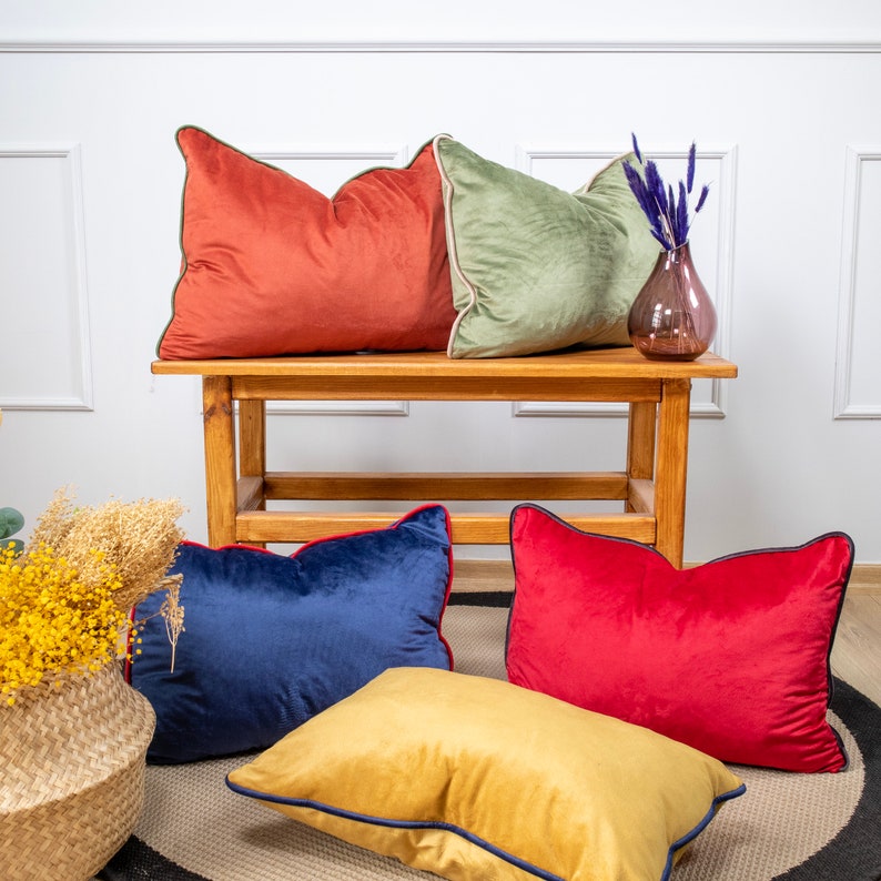 Pu&ograve; includere: Quattro cuscini decorativi di diversi colori e trame. I cuscini sono disposti su un tavolo di legno con un vaso di fiori secchi.