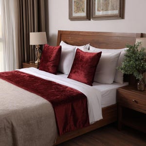 May include: A set of deep red velvet throw pillows and a matching velvet bed runner draped over a bed with white linens. The pillows are square and feature a soft, textured finish, while the long runner adds a bold color accent to the bedding.
