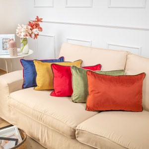 Pu&ograve; includere: Cinque cuscini decorativi su un divano color crema. I cuscini sono di vari colori, tra cui rosso, blu, giallo, verde e arancione. Ogni cuscino ha un bordo a contrasto attorno ai bordi.
