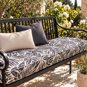 Puede incluir: Un banco de metal negro con un cojín de asiento largo con un patrón de hojas botánicas en blanco y negro. El banco incluye un cojín rectangular beige con textura de gofre y un cojín cuadrado gris oscuro con textura de gofre.