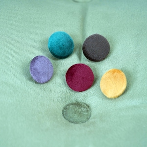 Puede incluir: Seis botones redondos de fieltro en varios colores: verde azulado, morado, burdeos, amarillo y dos tonos de gris. Los botones est&aacute;n dispuestos en semic&iacute;rculo sobre una superficie de tela verde.