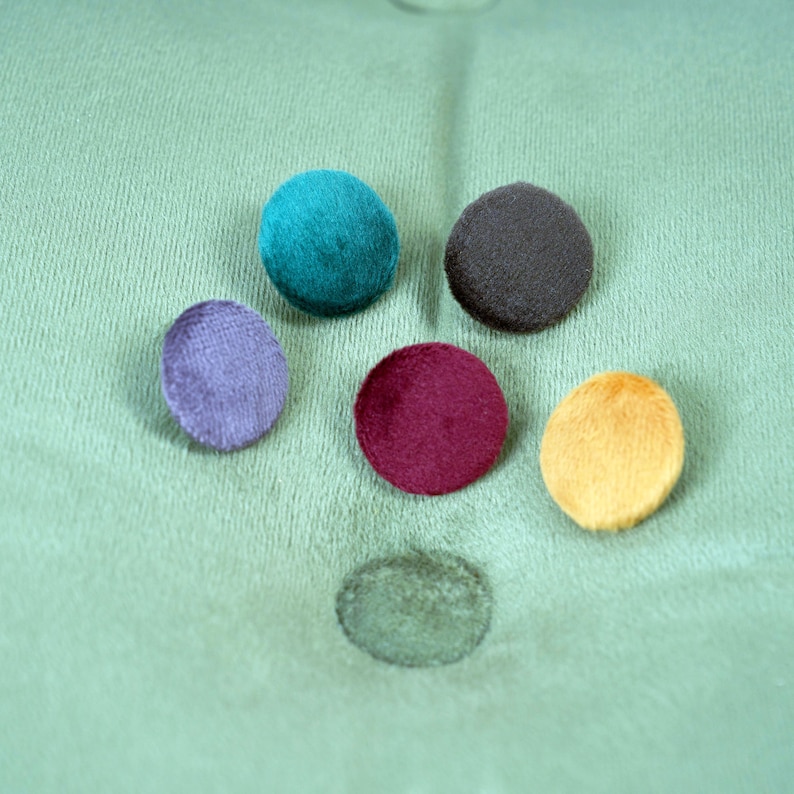 Puede incluir: Seis botones redondos de fieltro en varios colores: verde azulado, morado, burdeos, amarillo y dos tonos de gris. Los botones est&aacute;n dispuestos en semic&iacute;rculo sobre una superficie de tela verde.