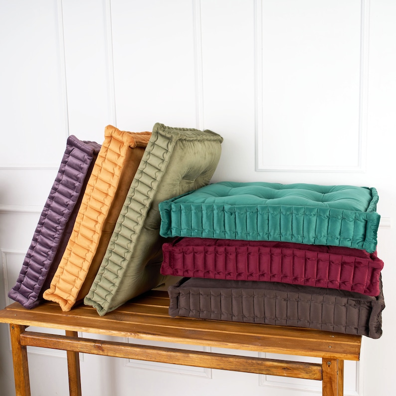 Puede incluir: Cinco cojines de suelo apilados en una mesa de madera. Los cojines son de terciopelo y tienen un dise&ntilde;o acolchado. Los cojines son en tonos de morado, naranja, verde, burdeos y marr&oacute;n.