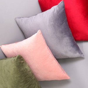 May include: Four decorative throw pillows in different colors and textures. The pillows are arranged in a stack, with the top pillow being red, followed by gray, pink, and green.