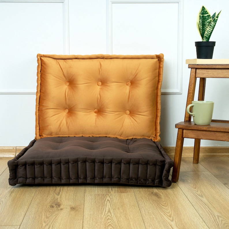 Puede incluir: Un coj&iacute;n de suelo marr&oacute;n y naranja con la parte superior acolchada. El coj&iacute;n est&aacute; hecho de tela y tiene una forma rectangular.