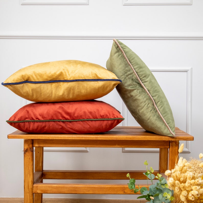 Pu&ograve; includere: Tre cuscini decorativi impilati su una panca di legno. Il cuscino superiore &egrave; verde scuro, il cuscino intermedio &egrave; arancione bruciato e il cuscino inferiore &egrave; giallo chiaro. Tutti e tre i cuscini hanno un bordo a contrasto.