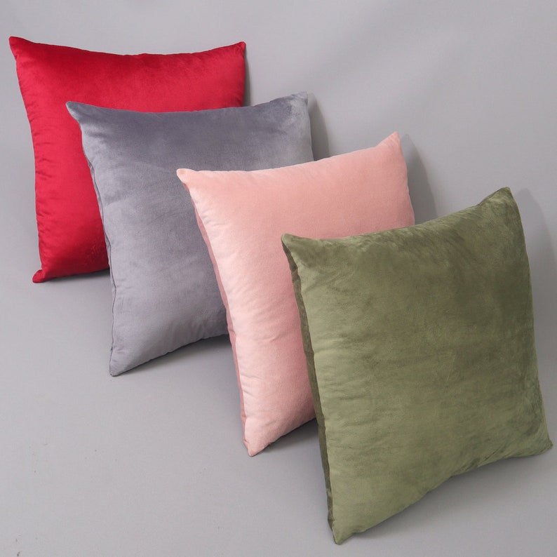May include: Four decorative throw pillows in different colors and textures. The pillows are arranged in a row, with the red pillow in the front, followed by gray, pink, and green.