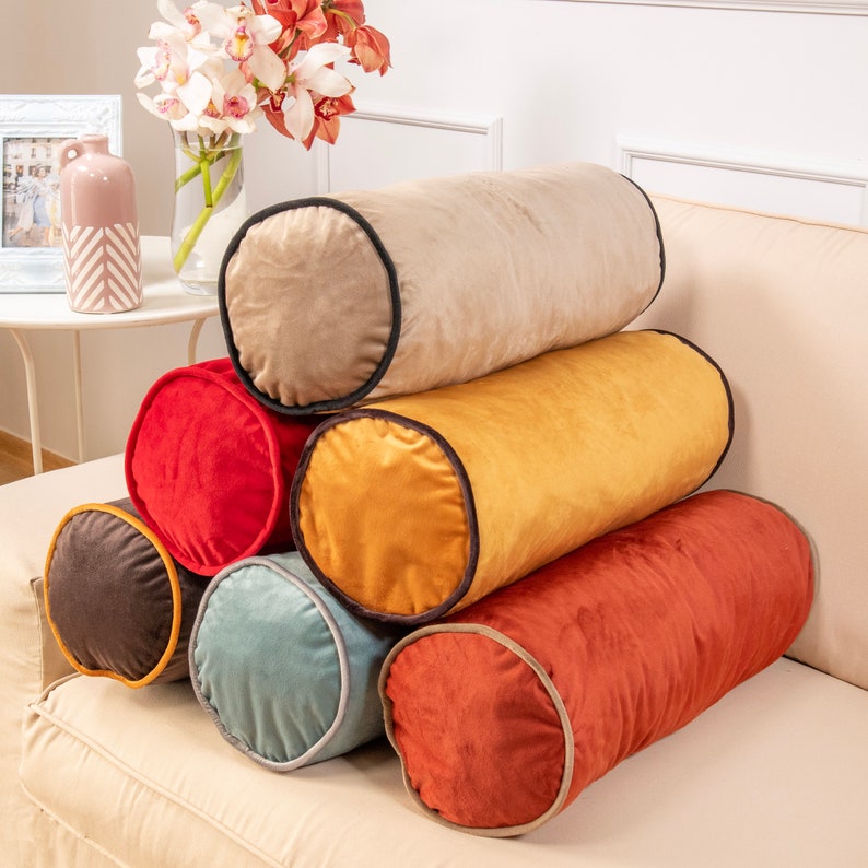 May include: A stack of six cylindrical throw pillows in various colours, including beige, red, yellow, brown, and blue. Each pillow has a black trim around the edges.