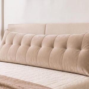 May include: Beige, tufted wedge pillow on a bed. The pillow has a rectangular shape with a series of vertical, buttoned sections. A pocket is visible on the side. The bed has a beige headboard and a white bedspread.