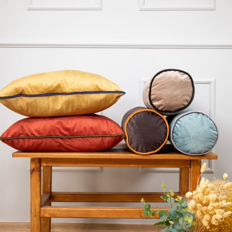 May include: Five decorative throw pillows in various colours and textures. The pillows are arranged on a wooden bench. The pillows include a mustard yellow pillow, a burnt orange pillow, a light brown pillow, a dark brown pillow, and a light blue pillow.