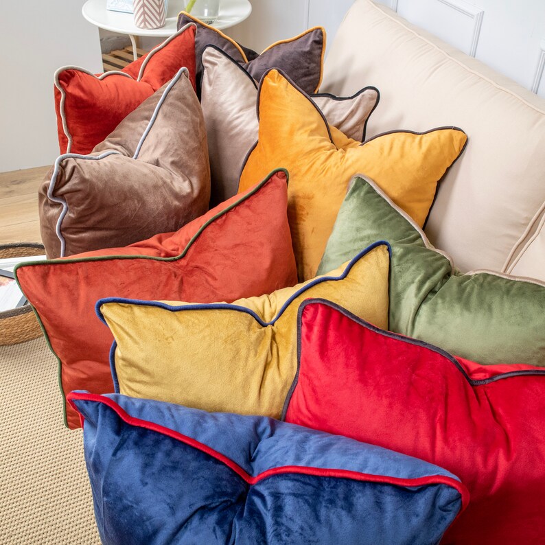 May include: A collection of decorative throw pillows with velvet fabric and contrasting trim. The pillows are in various colours, including brown, orange, yellow, green, red, and blue.