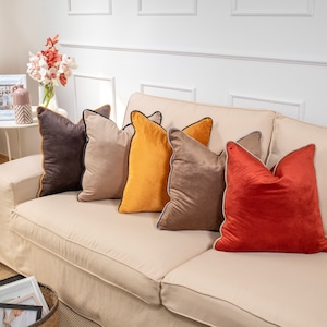 Pu&ograve; includere: Cinque cuscini decorativi in ​​velluto con bordatura a contrasto. I cuscini sono in tonalit&agrave; di marrone, beige, giallo e arancione. I cuscini sono disposti su un divano beige chiaro.