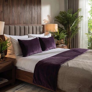 May include: A bedroom scene featuring a bed with white linens, two purple velvet throw pillows, and a matching bed runner. The wooden bed frame and headboard are complemented by a wooden nightstand and a large plant. The room has a warm, inviting atmosphere.