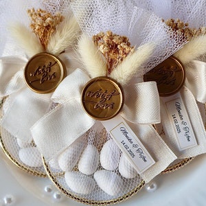 Puede incluir: Bolsitas de regalo blancas con sellos de cera dorados que dicen "with love" y una cinta blanca. Las bolsas contienen almendras blancas y tienen una pequeña etiqueta con los nombres "Monika & Tryfon" y la fecha "14.02.2025".