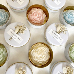 May include: A collection of floral-shaped candles in small, round, white tins with gold rims. The candles are various pastel colors, including pink, blue, and green. Each tin is topped with a white label that reads "Chama's Baby Shower 25.04.2023" and a small floral arrangement.