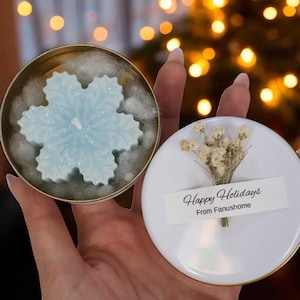 Puede incluir: Una vela azul claro en forma de copo de nieve en una lata dorada, junto a una tapa blanca con flores secas y el texto "Happy Holidays From Fanushome". Un árbol de Navidad iluminado se encuentra en el fondo.