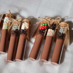 Puede incluir: Varios tubos de vidrio llenos de un polvo marrón y cubiertos con varios caramelos y chispas de colores. Cada tubo tiene un tapón de corcho atado con hilo y una pequeña etiqueta marrón. Los tubos están dispuestos sobre una superficie blanca.