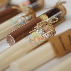 Puede incluir: Primer plano de varios tubos de vidrio llenos de varios ingredientes, incluyendo polvo marrón y chispas de colores. Cada tubo está sellado con un corcho y atado con hilo. Los tubos están dispuestos sobre una superficie blanca.