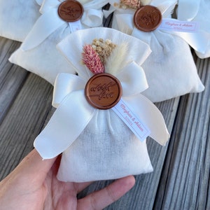 Puede incluir: Bolsas de tela blanca con un sello de cera marr&oacute;n y flores secas. Las bolsas tienen una cinta atada alrededor y una etiqueta que dice "Meghan & Adam 5.25.21".