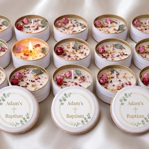 Può includere: Candele in scatola di latta bianche con decorazioni floreali e di foglie, alcune accese. Le candele sono decorate con petali di rosa secchi e verde. I coperchi di alcune candele hanno il testo "Adam's Baptism".