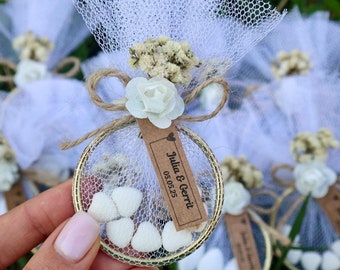 May include: White tulle favour bags with a small white flower and a gold circle containing white heart-shaped sweets. A brown tag with the text "Julia & Gerrit 05.05.23" is attached to the bag.