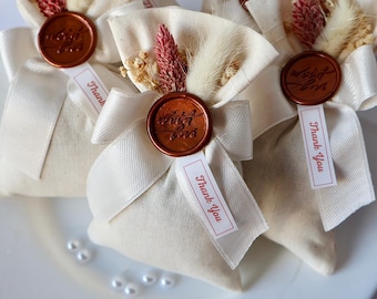 May include: Three small white fabric bags with a brown wax seal and a tag that says "Thank You". The bags are tied with a white ribbon and decorated with dried flowers.