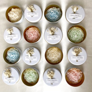 May include: An overhead shot of sixteen small, round, white tins with gold rims. Each tin contains a soap carved in the shape of a rose in various colors: white, blue, pink, and green. Each tin is decorated with a small bouquet and a label.