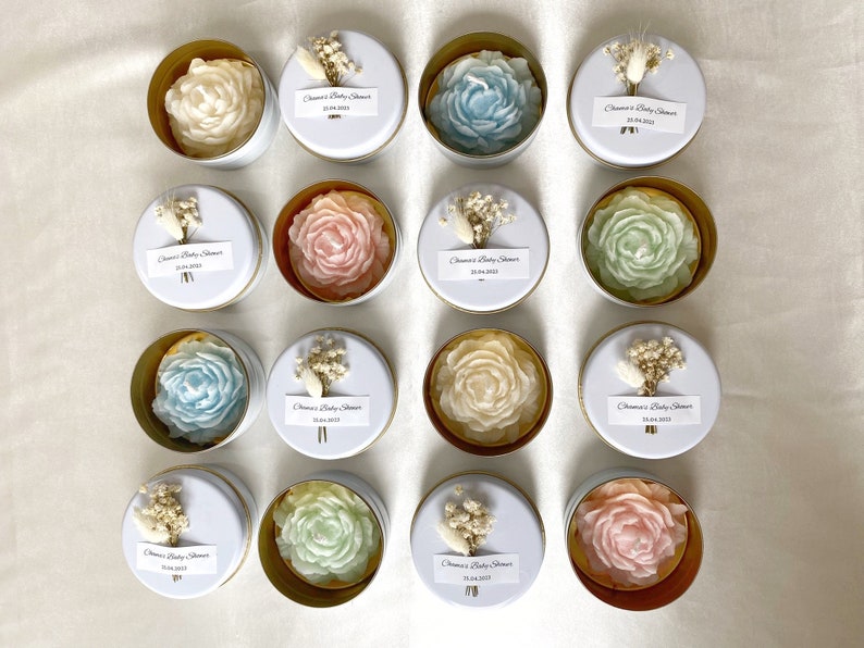 May include: An overhead shot of sixteen small, round, white tins with gold rims. Each tin contains a soap carved in the shape of a rose in various colors: white, blue, pink, and green. Each tin is decorated with a small bouquet and a label.