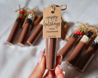 May include: A hand holds a clear glass tube filled with brown hot cocoa powder. A small brown tag reads "Happy holidays From Fanushome Hot Cocoa." Other tubes with decorative tops are in the background.
