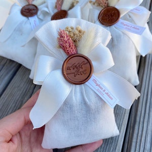Puede incluir: Bolsas de tela blanca con un sello de cera marr&oacute;n que dice "a little love". Las bolsas est&aacute;n atadas con una cinta de sat&eacute;n blanca y decoradas con flores secas. Una peque&ntilde;a etiqueta blanca con el texto "Meghan & Adam 5.18.24" est&aacute; unida a una de las bolsas.
