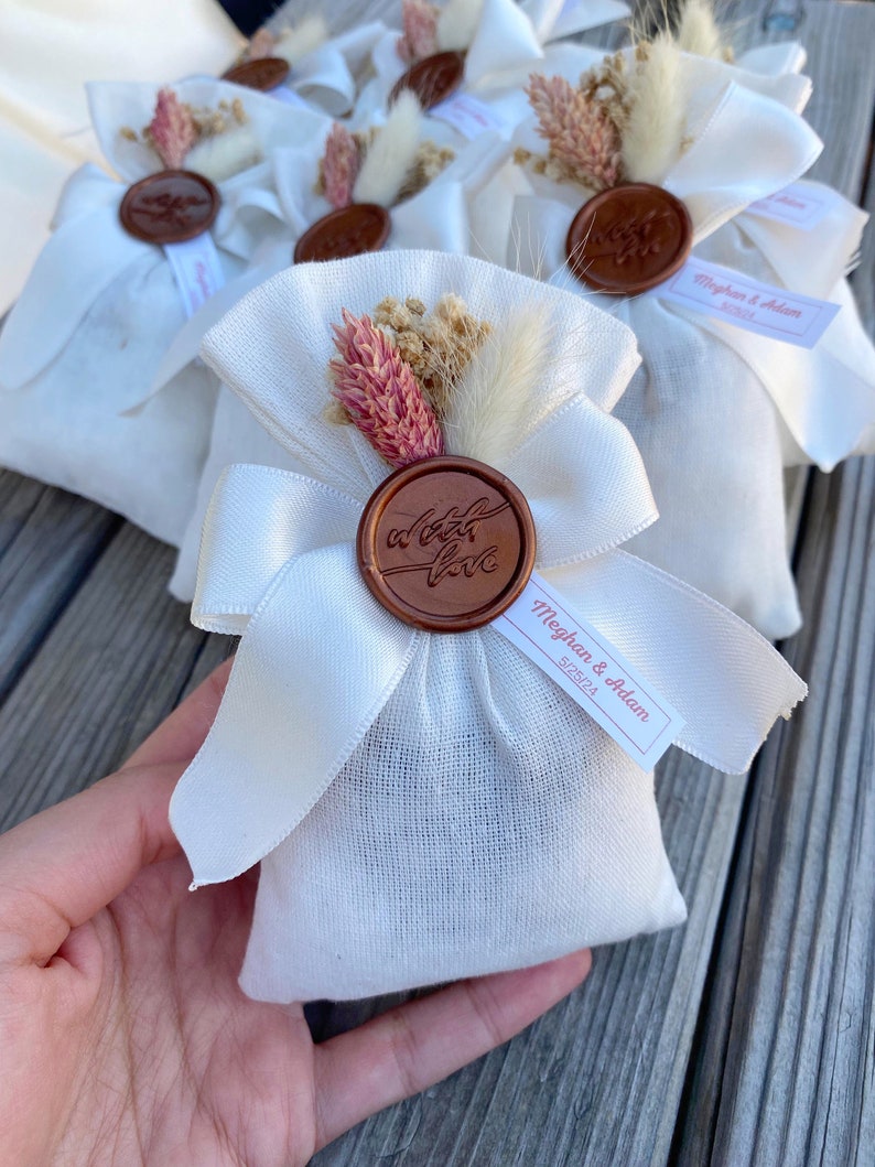 Puede incluir: Bolsas de tela blanca con un sello de cera marr&oacute;n que dice "a little love". Las bolsas est&aacute;n atadas con una cinta de sat&eacute;n blanca y decoradas con flores secas. Una peque&ntilde;a etiqueta blanca con el texto "Meghan & Adam 5.18.24" est&aacute; unida a una de las bolsas.